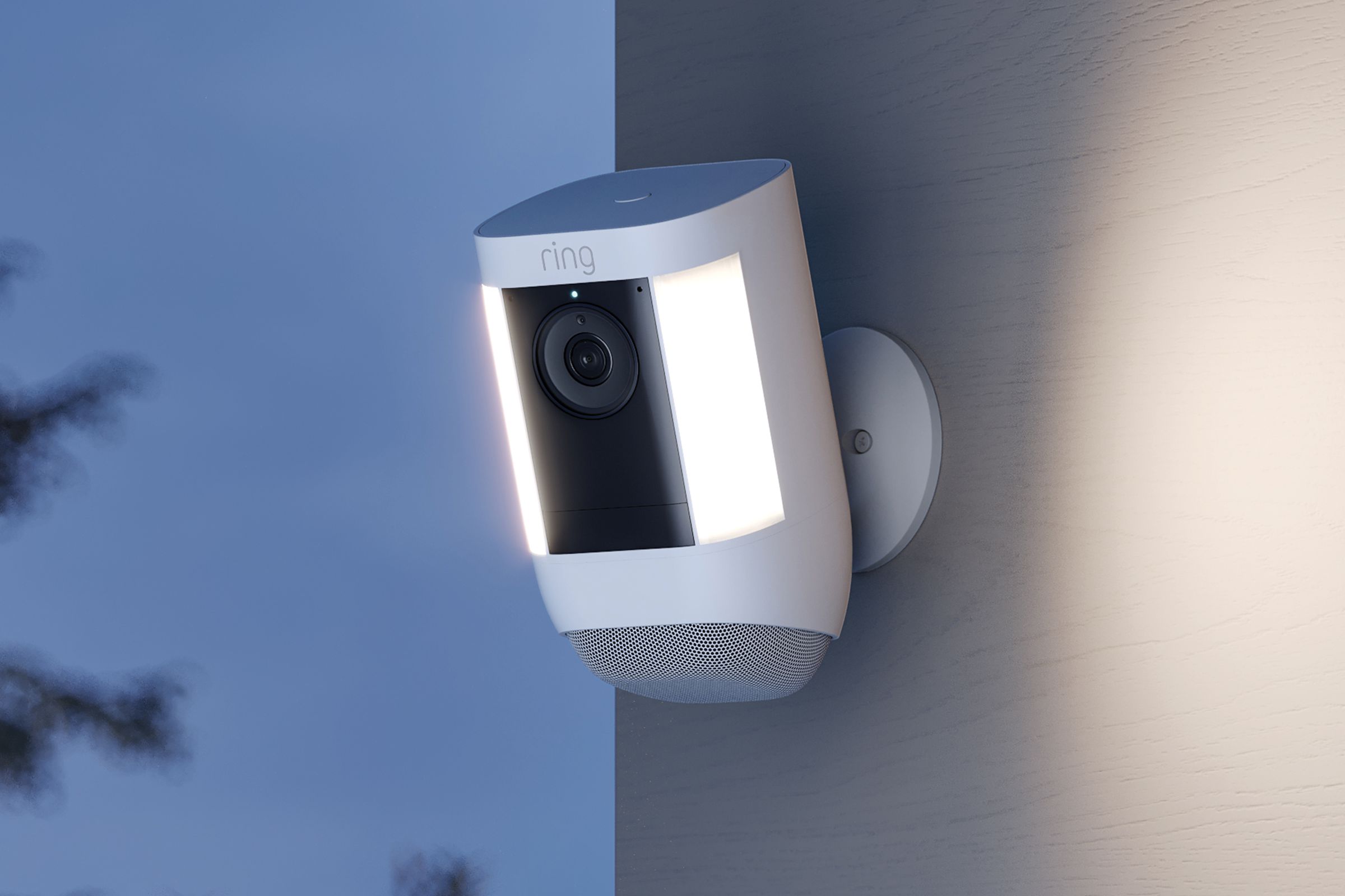 Ring’s spotlight camera is installed on an outdoor. Off the wall is a dark wooded background, and the camera has two lights on either side of its body on.