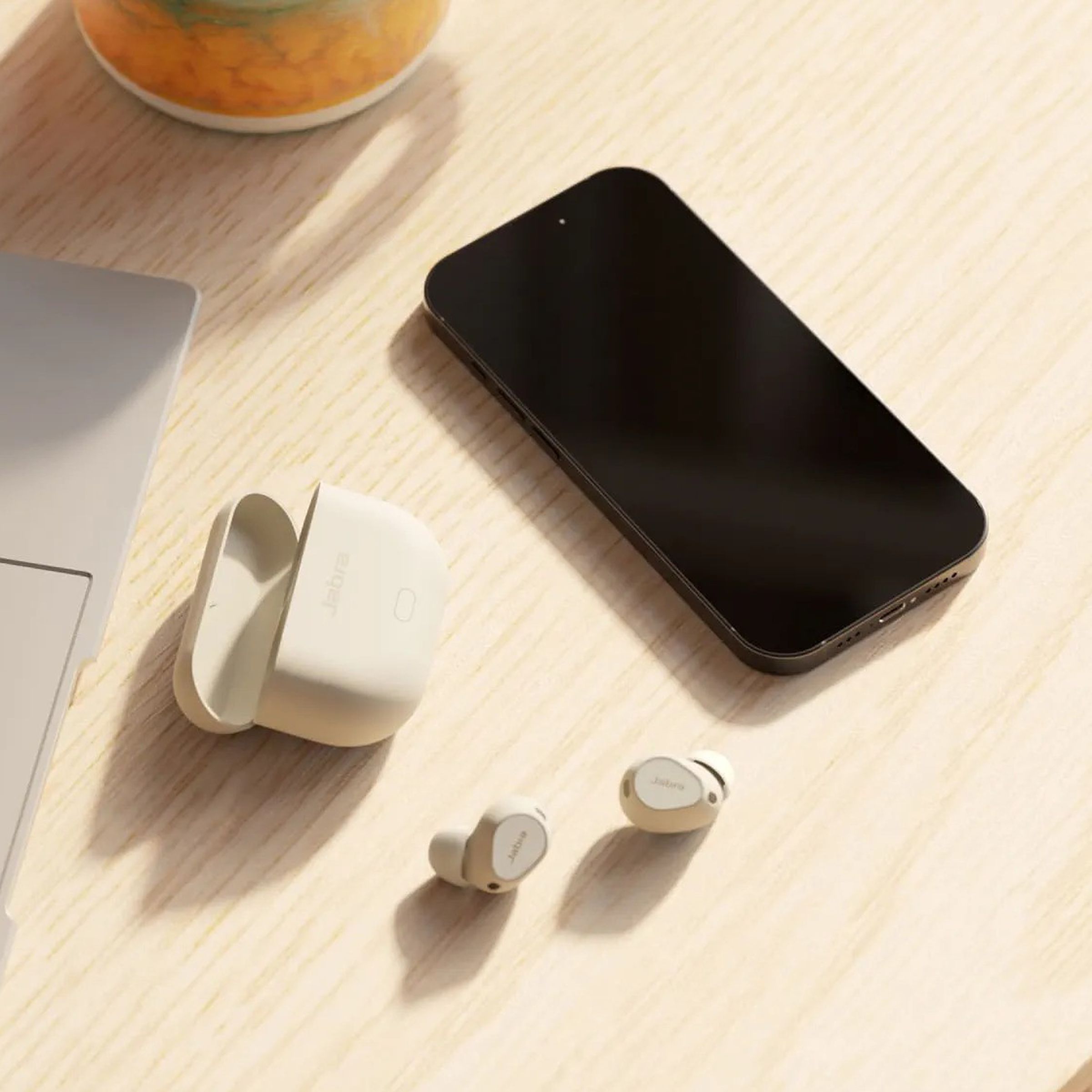 The Jabra Elite 10 gen 2 wireless earbuds on a table besides a laptop and a smartphone.