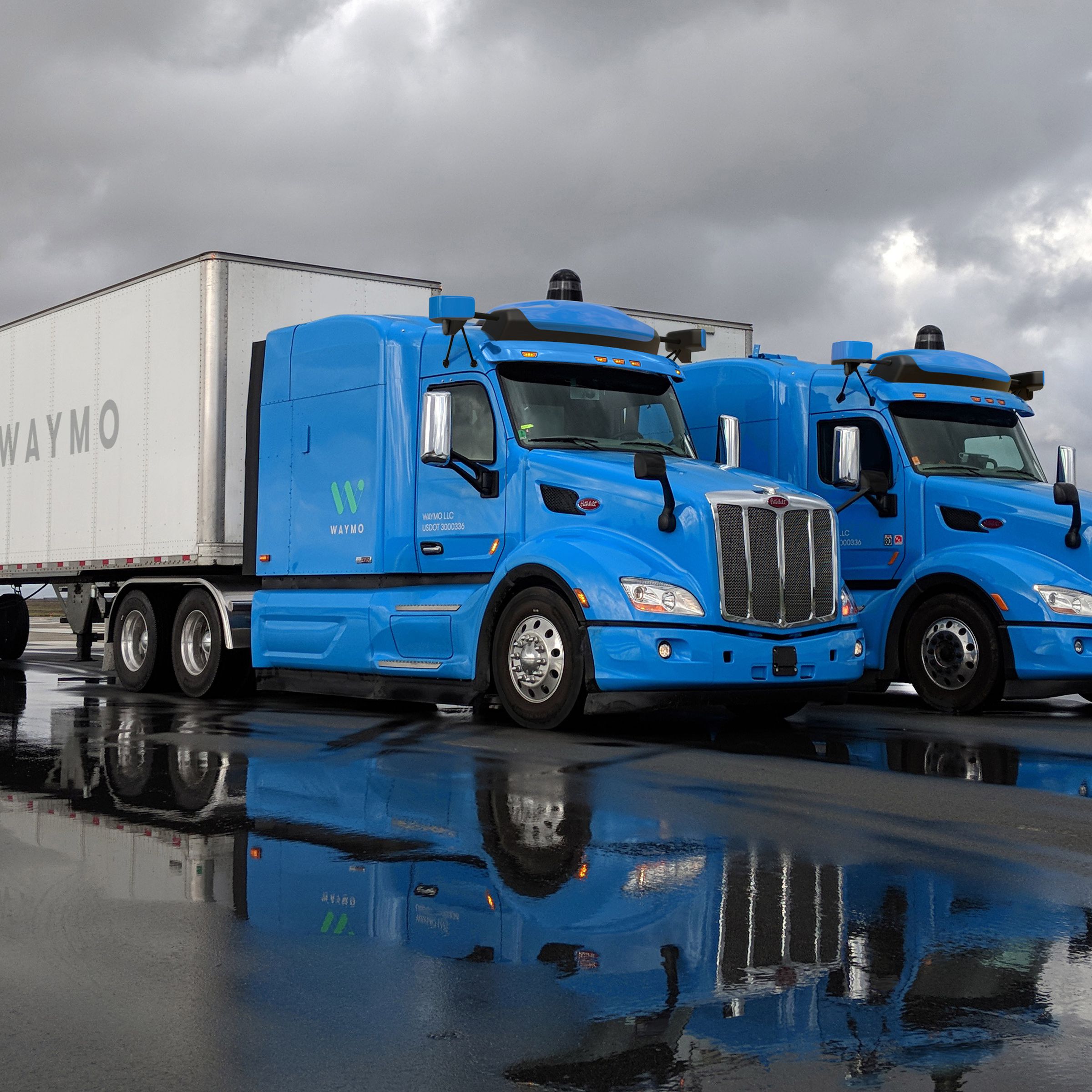 Two blue Waymo autonomous trucks next to one another on a cloudy, rainy day.