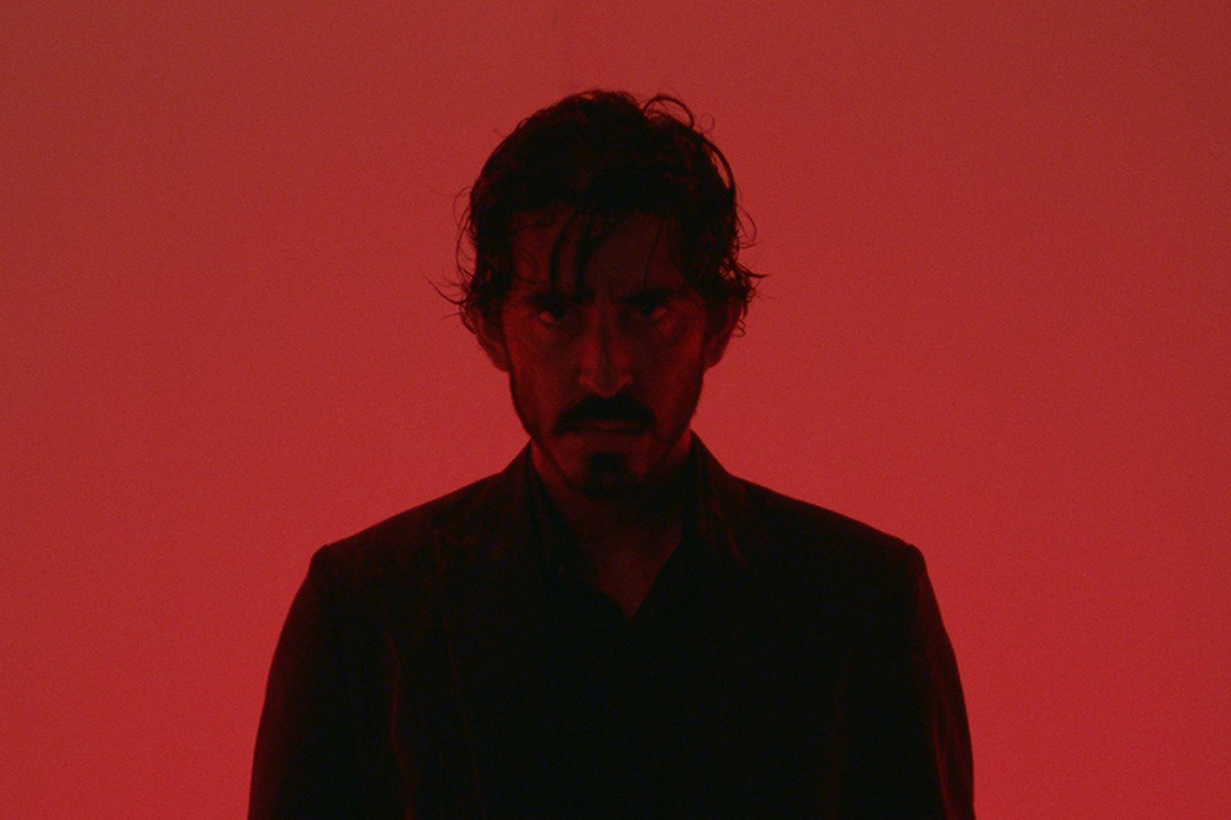 A man in a black suit and shirt backlit by red lighting that casts him in shadow.